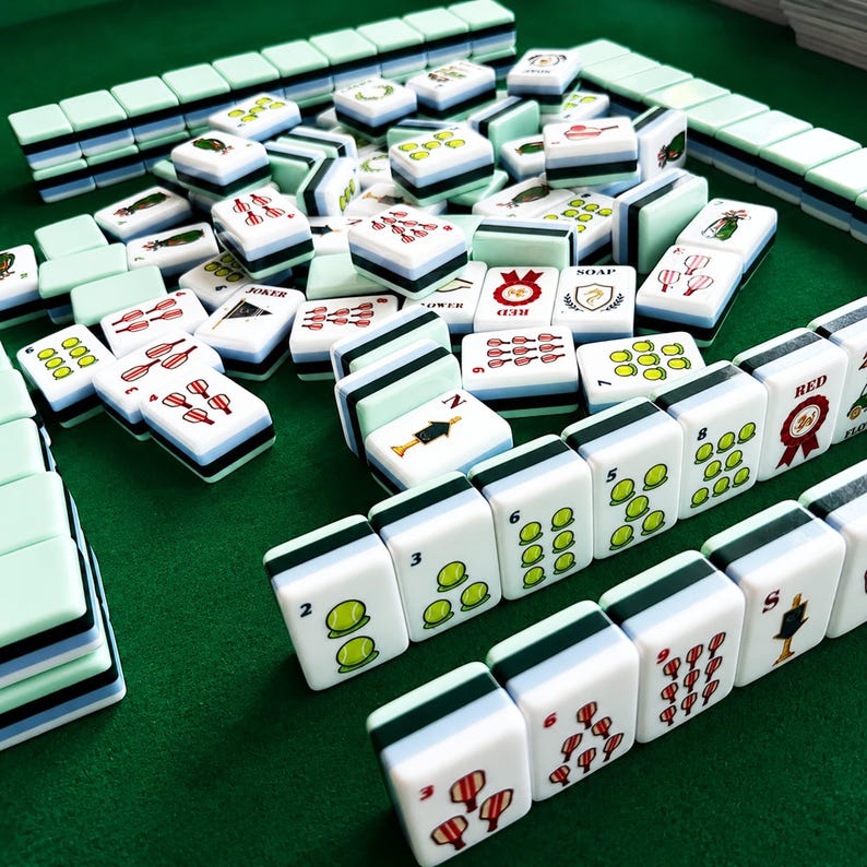 Pode incluir: Um conjunto de pe&ccedil;as retangulares de mahjong com faces brancas e lados em camadas nas cores verde-claro, azul e preto. As pe&ccedil;as apresentam v&aacute;rias ilustra&ccedil;&otilde;es coloridas, incluindo bolas de t&eacute;nis, fitas vermelhas e texto como RED, SOAP e JOKER numa superf&iacute;cie verde.