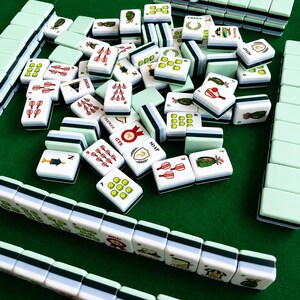 Pode incluir: Um conjunto de pe&ccedil;as retangulares de mahjong com faces brancas e versos verde-claros. As pe&ccedil;as apresentam v&aacute;rias ilustra&ccedil;&otilde;es coloridas, incluindo tacos de golfe, bolas e etiquetas de texto como "JOKER", "FLOWER", "RED", "GREEN" e "SOAP".