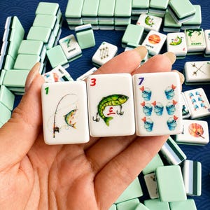 May include: A close-up of a Mahjong game set. Three tiles are held in the foreground, displaying colorful illustrations of a fish, fishing rod, and buckets. The tiles are white with green and black accents. Other tiles are stacked in the background.