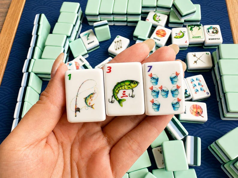 May include: A close-up of a Mahjong game set. Three tiles are held in the foreground, displaying colorful illustrations of a fish, fishing rod, and buckets. The tiles are white with green and black accents. Other tiles are stacked in the background.