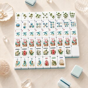 May include: A set of white Mahjong tiles with various illustrations, including palm trees, seashells, and flamingos. The tiles are arranged on a light surface, with small blue boxes and seashells nearby. The tiles feature numbers, suits, and seasonal themes.
