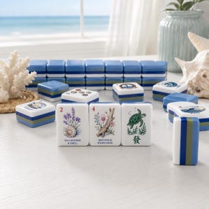 May include: A set of rectangular mahjong tiles featuring coastal-themed illustrations, including sea lavender, shells, driftwood, wildflowers, and a sea turtle. The tiles have white faces with colorful graphics and blue, green, and white striped sides.
