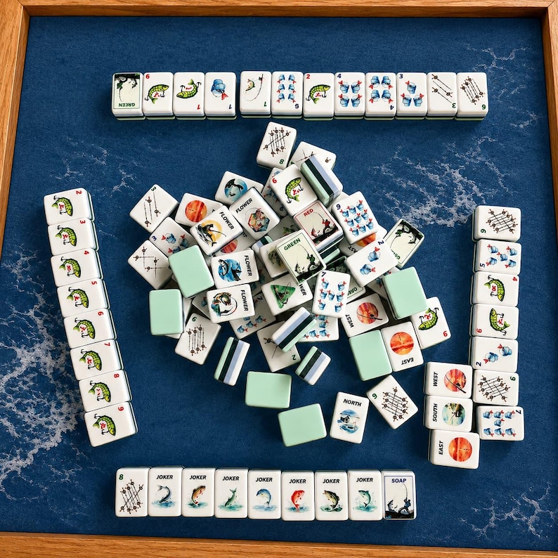 May include: A collection of white Mahjong tiles with various symbols and characters, including flowers, fish, and directional markers. The tiles are arranged on a blue playing surface, with some stacked and others scattered.