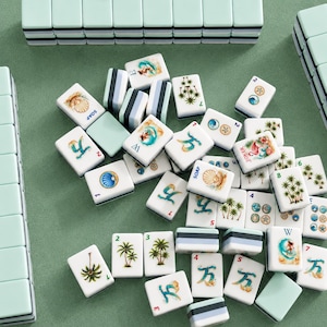 May include: A collection of white Mahjong tiles with various illustrations, including palm trees, shells, and figures. The tiles are arranged on a green surface, with some tiles stacked and others scattered. The set includes tiles with the word "SOAP" and numbers.