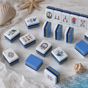 May include: A collection of rectangular mahjong tiles featuring nautical and coastal illustrations, including lighthouses, anchors, sea creatures, and compasses. The tiles have white faces with blue and green layered bases and some include text.