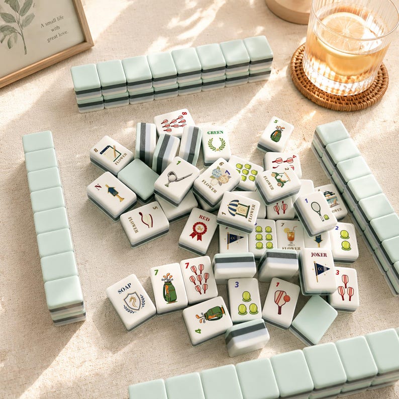 May include: A collection of Mahjong tiles in a variety of designs and colors, including green, white, and gray. The tiles are arranged on a light-colored surface, with some stacked and others scattered. A glass of water with a lemon slice is also visible.