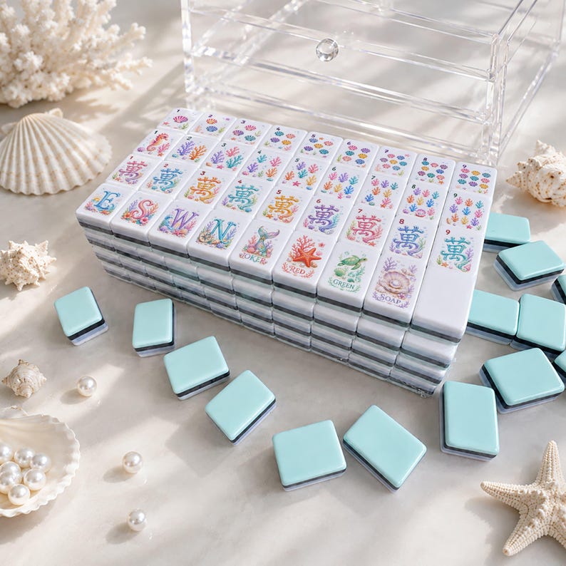 Op de afbeelding: Een set witte Mahjong-tegels met kleurrijke illustraties van zeeleven en Chinese karakters. De tegels zijn gestapeld, met extra lichtblauwe tegels verspreid. Een transparante acryl opbergdoos staat op de achtergrond, samen met schelpen en parels.