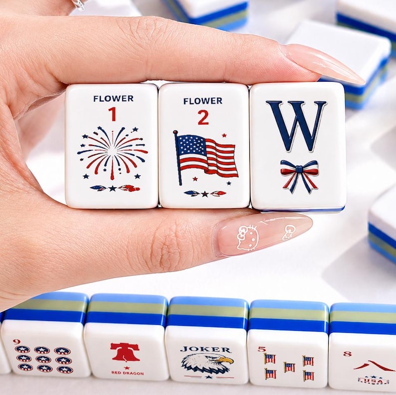 May include: White Mahjong tiles with blue trim, featuring red, white, and blue designs. The tiles include a firework, an American flag, and the letter "W" with a bow. Other tiles show various symbols and numbers.