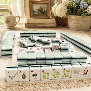 May include: A collection of white and green Mahjong tiles with various symbols and numbers. The tiles are arranged on a light-colored surface, with some stacked and others scattered. The image includes a framed picture, books, and floral arrangements.
