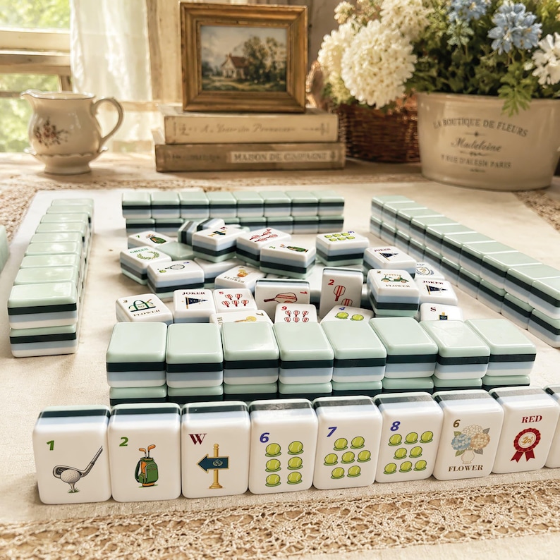 May include: A collection of white and green Mahjong tiles with various symbols and numbers. The tiles are arranged on a light-colored surface, with some stacked and others scattered. The image includes a framed picture, books, and floral arrangements.