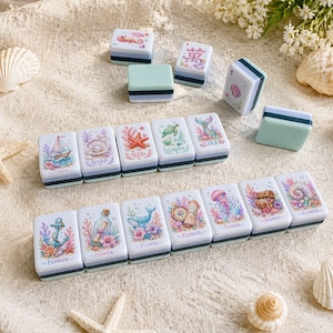 Op de afbeelding: Een verzameling decoratieve, rechthoekige zeepbakjes met mariene illustraties. Elk bakje heeft een uniek ontwerp, waaronder een zeilboot, zeester en kwal, met een witte basis, een zwarte streep en een lichtblauwe bovenkant. De bakjes zijn op een zandige ondergrond geplaatst.