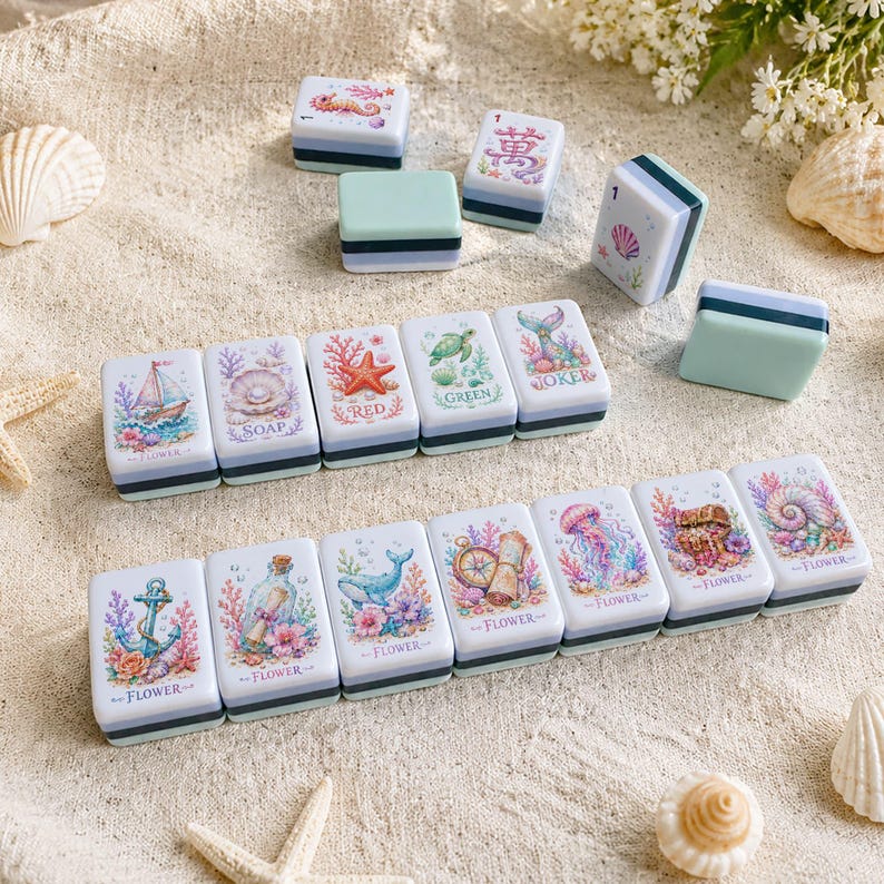 Op de afbeelding: Een verzameling decoratieve, rechthoekige zeepbakjes met mariene illustraties. Elk bakje heeft een uniek ontwerp, waaronder een zeilboot, zeester en kwal, met een witte basis, een zwarte streep en een lichtblauwe bovenkant. De bakjes zijn op een zandige ondergrond geplaatst.