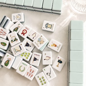 May include: A collection of white Mahjong tiles with various illustrations and numbers, including golf-themed images, flowers, and the word "SOAP". The tiles are arranged on a light surface, with stacks of light green tiles in the background.