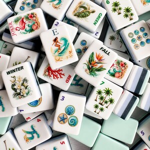May include: A collection of white Mahjong tiles with colorful illustrations. Some tiles feature tropical scenes, a flamingo, a surfer, and floral designs. Other tiles have numbers and words like "Joker", "Winter", and "Fall".
