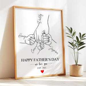 Könnte beinhalten: Ein gerahmter Druck mit einer minimalistischen Strichzeichnung von Händen, die sich halten. Der Druck enthält die Worte "Happy Father's Day, we love you, Est. 2021" und die Namen "Henry, Cora, Jasmine, Daddy". Unten befindet sich ein rotes Herz.