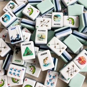 May include: A collection of white and seafoam green Mahjong tiles. The tiles feature fishing-themed illustrations, including fish, fishing rods, and boats. Some tiles have the word "FLOWER" or numbers.