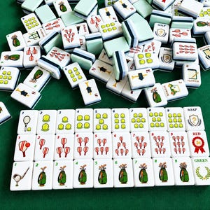 Pode incluir: Um conjunto de pe&ccedil;as brancas de mahjong com ilustra&ccedil;&otilde;es de tem&aacute;tica desportiva, incluindo bolas de t&eacute;nis, tacos de golfe e raquetes. As pe&ccedil;as est&atilde;o dispostas em filas com etiquetas numeradas e texto como SOAP, RED e GREEN.