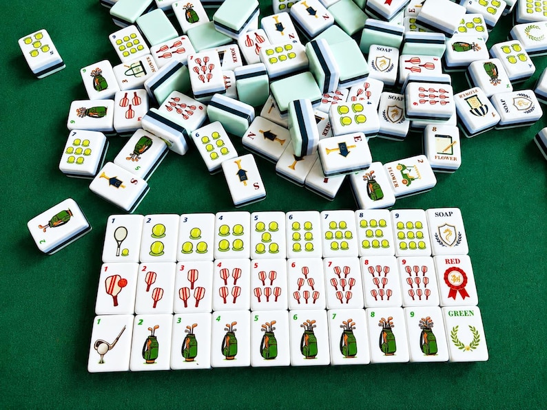 Pode incluir: Um conjunto de pe&ccedil;as brancas de mahjong com ilustra&ccedil;&otilde;es de tem&aacute;tica desportiva, incluindo bolas de t&eacute;nis, tacos de golfe e raquetes. As pe&ccedil;as est&atilde;o dispostas em filas com etiquetas numeradas e texto como SOAP, RED e GREEN.