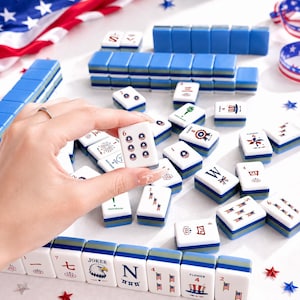 May include: A complete Mahjong set with blue and white tiles, some featuring patriotic designs and the number 6. The set includes blue racks and is arranged on a white surface with American flag decorations.