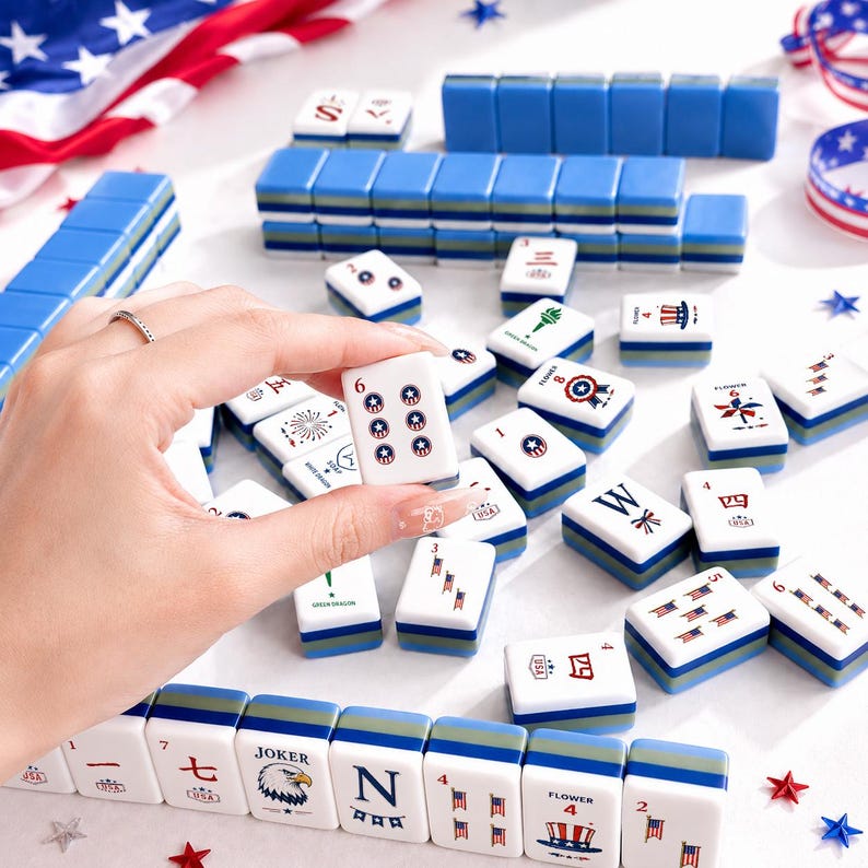 May include: A complete Mahjong set with blue and white tiles, some featuring patriotic designs and the number 6. The set includes blue racks and is arranged on a white surface with American flag decorations.