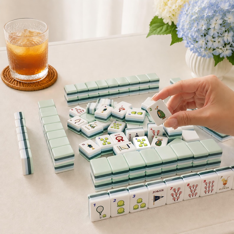 May include: A collection of white and green Mahjong tiles arranged on a white surface. The tiles feature various symbols and numbers. A hand holds a tile with the number 2 and a golf bag symbol. A glass of iced tea sits on a woven coaster.