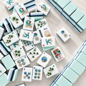 May include: A collection of white Mahjong tiles with various beach-themed illustrations, including palm trees, seashells, and surfers. The tiles have a light blue and white border. Some tiles have numbers and the word "SOAP" printed on them.