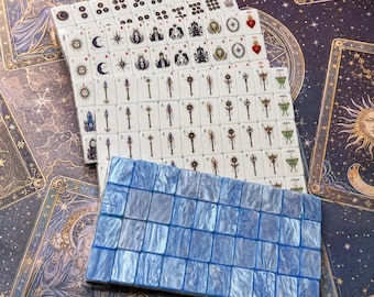 May include: A set of rectangular mahjong tiles featuring white tiles with black and colored illustrations of celestial bodies, wands, and hearts, alongside a row of solid, pearlescent blue tiles. The tiles display various symbols, numbers, and icons.