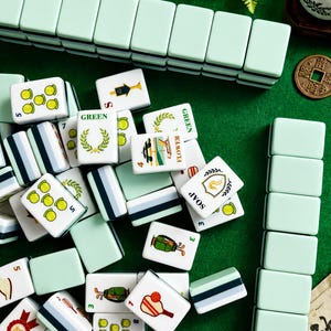 Pode incluir: Um conjunto de pe&ccedil;as retangulares de mahjong com versos em verde menta claro e faces brancas apresentando v&aacute;rias ilustra&ccedil;&otilde;es coloridas, incluindo equipamentos esportivos, coroas de flores e etiquetas de texto como GREEN, FLOWER, SOAP e RED.