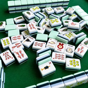 Pode incluir: Um conjunto de pe&ccedil;as de Mahjong com verso verde menta claro e frente branca com v&aacute;rias ilustra&ccedil;&otilde;es coloridas, incluindo n&uacute;meros, s&iacute;mbolos e texto como JOKER, FLOWER, RED e SOAP, dispostas em uma pilha e em fileiras organizadas.