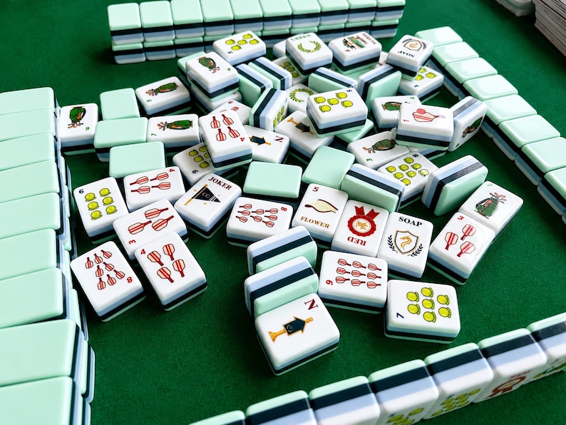 Pode incluir: Um conjunto de pe&ccedil;as de Mahjong com verso verde menta claro e frente branca com v&aacute;rias ilustra&ccedil;&otilde;es coloridas, incluindo n&uacute;meros, s&iacute;mbolos e texto como JOKER, FLOWER, RED e SOAP, dispostas em uma pilha e em fileiras organizadas.
