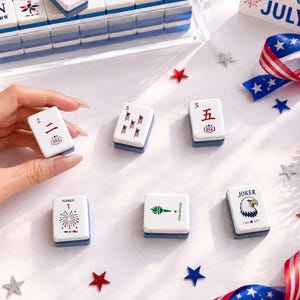 May include: A collection of white and blue Mahjong tiles with various symbols and numbers, including a "Joker" tile with an eagle. The tiles are arranged with patriotic decorations like star-shaped confetti and a ribbon with stars.
