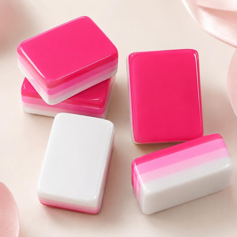 May include: A collection of rectangular mahjong tiles featuring a layered gradient design. The tiles transition from a solid hot pink top layer through lighter pink and white bands. Some tiles are stacked, while others lie flat on the surface.
