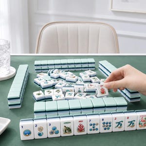 May include: A Mahjong set with white and teal tiles is arranged on a green surface. The tiles feature various symbols and numbers. A hand is picking up a tile with a red coral design. A glass of water sits on the left.