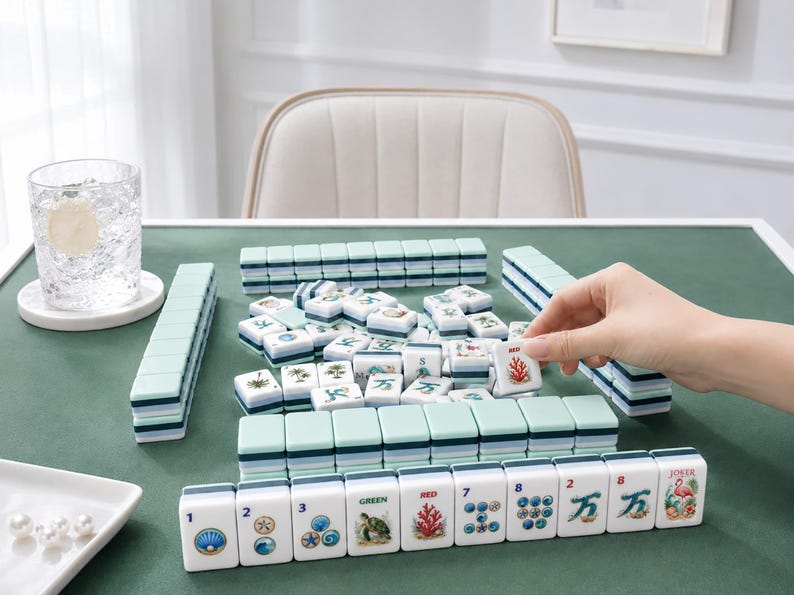 May include: A Mahjong set with white and teal tiles is arranged on a green surface. The tiles feature various symbols and numbers. A hand is picking up a tile with a red coral design. A glass of water sits on the left.
