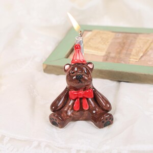 Peut inclure: Une figurine de bougeoir en forme d'ours brun en céramique avec une finition brillante, un chapeau de fête rouge et un nœud papillon rouge. Une bougie argentée allumée est insérée sur le dessus de la tête de l'ours, émettant une flamme.
