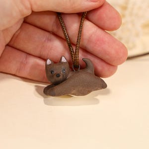 May include: A brown ceramic dog pendant on a brown cord. The dog is lying down with its ears perked up. The pendant is held by a hand against a neutral background. The dog has black eyes and pink accents on its ears.