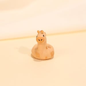 Könnte beinhalten: Kleine, hellbraune Giraffenfigur mit braunen Flecken. Die Giraffe hat einen runden Körper und einen langen Hals mit kleinen Ohren. Die Figur besteht aus einem glatten Material und steht vor einem hellen Hintergrund.