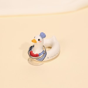 Pode incluir: Um porta-anéis em forma de pato de cerâmica branca, com chapéu e gola de marinheiro azuis e laço vermelho. O pato tem um bico amarelo e foi concebido para segurar anéis. Um artigo caprichoso e decorativo.