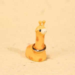 Peut inclure: Un porte-bague girafe fantaisiste. La figurine est jaune avec des taches marron et un museau blanc. Une bague argentée est présentée autour du cou de la girafe. Le porte-bague est un objet décoratif pour bijoux.