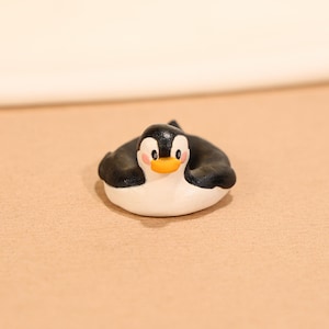 May include: A small, handcrafted penguin figurine. The penguin has a black head and wings, a white body, and an orange beak. The cheeks are pink. The figurine is resting on a tan surface.