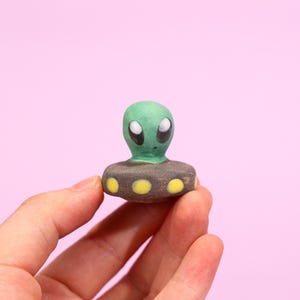 Puede incluir: Una pequeña figura de alienígena pintada a mano, con cabeza verde y grandes ojos negros. El alienígena está sobre una base gris oscuro decorada con puntos amarillos. El fondo es rosa liso.