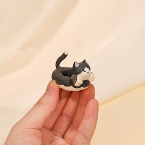 Könnte beinhalten: Ein kleiner, keramischer Katzen-Pflanztopf. Die Katze ist schwarz und weiß mit einem eingerollten Schwanz und hat die Form eines Donuts. Der Pflanztopf wird in einer Hand vor einem neutralen Hintergrund gehalten. Der Katzen-Pflanztopf ist ein dekorativer Artikel.