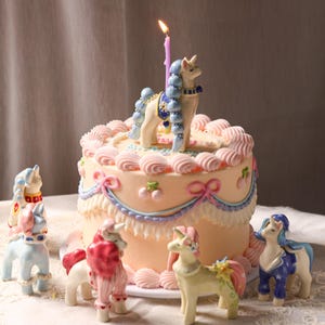 May include: A decorated cake with a lit candle and several unicorn figurines. The cake is light orange with pink frosting, bows, and a blue and pink trim. The unicorns are various colors, including blue, pink, and yellow.