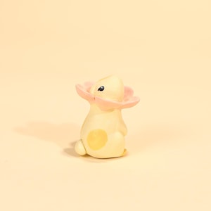 Puede incluir: Una figura caprichosa pintada a mano de un conejito con una flor rosa alrededor de la cabeza. El conejito es de color amarillo claro con una mancha amarilla en la espalda. La figura está sobre un fondo amarillo suave.