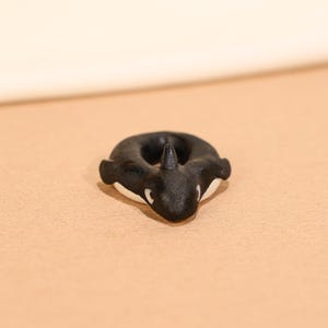 Op de afbeelding: Een kleine, zwart-witte orka-ring. De ring heeft de vorm van een donut met een rugvin en witte markeringen op de buik en ogen. De ring is gemaakt van een glad materiaal en ligt op een bruinachtige ondergrond.