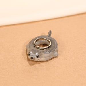Puede incluir: Un soporte para anillos de foca de cerámica gris con un anillo plateado. La foca tiene ojos negros y una pequeña cola. El anillo es una banda plateada con un diseño trenzado. El soporte para anillos está sobre una superficie beige.