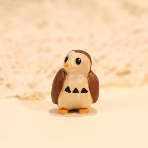 May include: A small, handcrafted owl figurine. The owl has a light brown body with dark brown wings and head. It features large, round eyes with yellow irises and black pupils. Three black triangles adorn the chest.