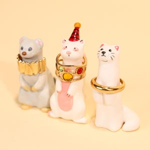 May include: Three ceramic ferret figurines, each with a ring around its neck. One gray ferret has a gold collar, another white ferret wears a ring with colorful gems, and the third white ferret has a simple gold ring.