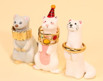 May include: Three ceramic ferret figurines, each with a ring around its neck. One gray ferret has a gold collar, another white ferret wears a ring with colorful gems, and the third white ferret has a simple gold ring.