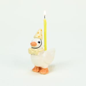 Könnte beinhalten: Ein Kerzenhalter für den Geburtstag in Form einer Gans. Die Gans ist weiß mit orangefarbenen Füßen und Schnabel, trägt eine gelbe Fliege und einen Partyhut mit gelben Punkten. Eine brennende gelbe Kerze ist in den Rücken der Gans gesteckt.